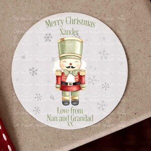 May include: A wrapped gift box with a burgundy ribbon and a round sticker. The sticker features a nutcracker illustration and the text "Merry Christmas Xander". It also includes the text "Love from Nan and Grandad".