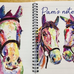 May include: A notebook featuring a colourful watercolour painting of three horses on the cover. The horses are wearing bridles and have a variety of colours, including pink, blue, yellow, and green. The notebook is titled "Pam's notes".