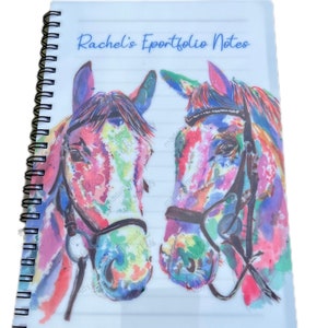 May include: A white spiral bound notebook with a clear plastic cover. The cover features a watercolour painting of two colourful horse heads with bridles. The text "Rachel's Eportfolio Notes" is printed in blue on the cover.