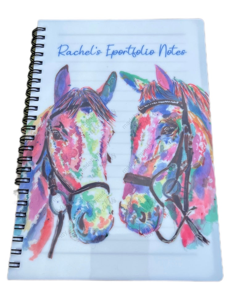 May include: A white spiral bound notebook with a clear plastic cover. The cover features a watercolour painting of two colourful horse heads with bridles. The text "Rachel's Eportfolio Notes" is printed in blue on the cover.