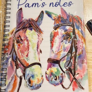 May include: A white notebook with a spiral binding. The cover features a watercolour painting of two horses' heads with colourful details. The text "Pam's notes" is printed in blue on the cover.