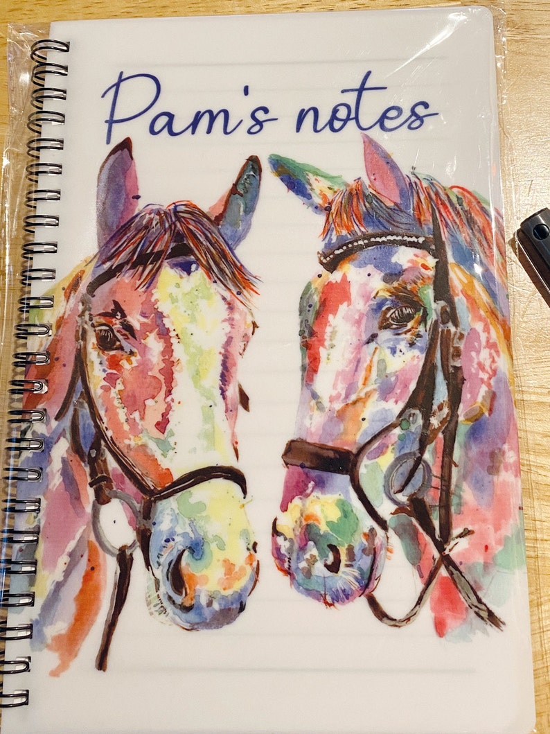 May include: A white notebook with a spiral binding. The cover features a watercolour painting of two horses' heads with colourful details. The text "Pam's notes" is printed in blue on the cover.