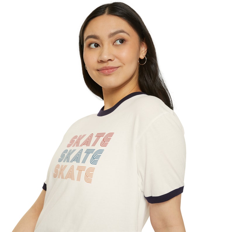 Op de afbeelding: Een wit T-shirt met een marineblauwe boord en mouwen. Het shirt heeft een grafische print van het woord "SKATE" dat drie keer in verschillende kleuren wordt herhaald.