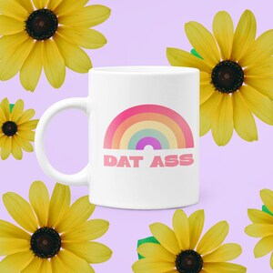 May include: White ceramic mug featuring a rainbow design and the text "DAT ASS" in pink. The mug is surrounded by yellow sunflowers on a lavender background. A fun and colorful gift idea.