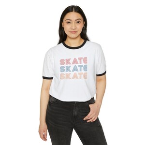 Op de afbeelding: Wit T-shirt met een zwarte boord en het woord "SKATE" drie keer herhaald in een retro-lettertype met roze, blauwe en oranje strepen.