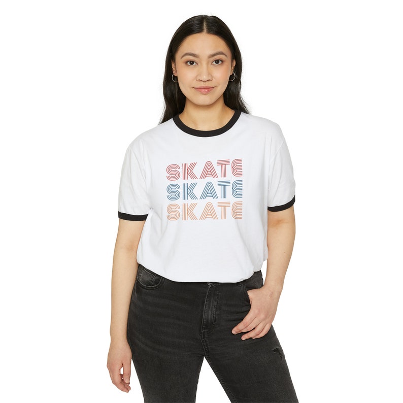 Op de afbeelding: Wit T-shirt met een zwarte boord en het woord "SKATE" drie keer herhaald in een retro-lettertype met roze, blauwe en oranje strepen.