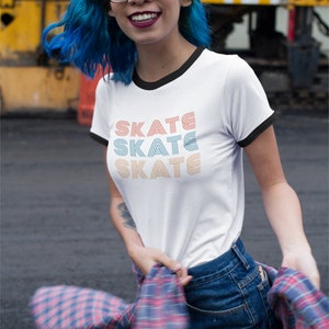 Op de afbeelding: Een wit T-shirt met een zwarte boord en het woord "SKATE" drie keer herhaald in een retro-lettertype. De tekst is in oranje, blauw en lichtblauw. De persoon die het shirt draagt heeft blauw haar en draagt een geruit overhemd om de taille gebonden.