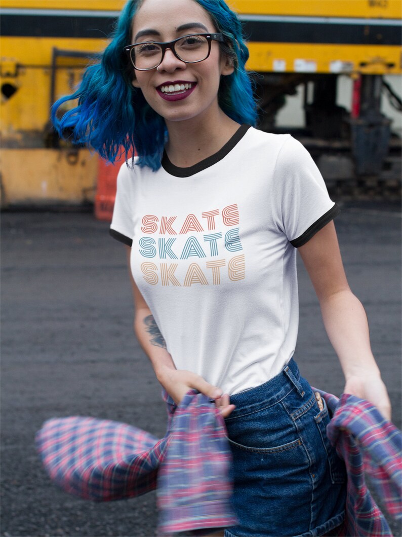 Op de afbeelding: Een wit T-shirt met een zwarte boord en het woord "SKATE" drie keer herhaald in een retro-lettertype. De tekst is in oranje, blauw en lichtblauw. De persoon die het shirt draagt heeft blauw haar en draagt een geruit overhemd om de taille gebonden.