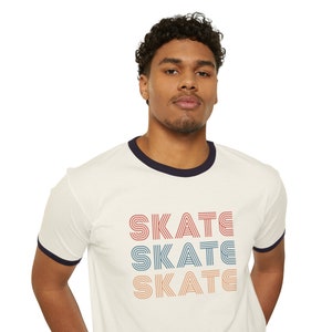 Op de afbeelding: Een wit T-shirt met een marineblauwe boord. Het shirt heeft een retro-stijl grafische afbeelding van het woord "SKATE" dat drie keer in verschillende kleuren wordt herhaald.