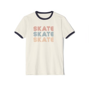Op de afbeelding: Een wit T-shirt met een marineblauwe boord. Het shirt heeft een grafische afbeelding van het woord "SKATE" dat drie keer wordt herhaald in een retro-lettertype met de kleuren rood, oranje en blauw.