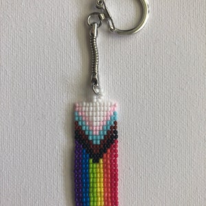 May include: A beaded keychain featuring a rectangular design with a rainbow gradient and a transgender pride flag. The keychain has a silver-colored metal ring and a spring-like connector. The beaded section is approximately 1.5 inches long.