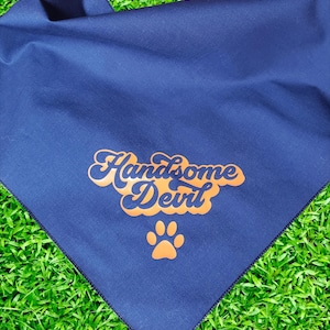 Puede incluir: Un bandana azul marino con el texto "Handsome Devil" en letra naranja y una huella de pata naranja.
