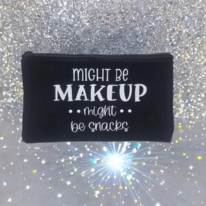 May include: Black zippered cosmetic bag with the text "Might Be Makeup ... might ... be snacks" in white. The bag is rectangular and appears to be made of a soft material. The background is a sparkly, glittery surface.
