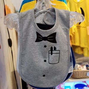 May include: A gray baby bib with a black bow tie graphic, a pocket design, and two small pen or pencil graphics. The bib has a rounded bottom and is hanging on a clear plastic hanger.
