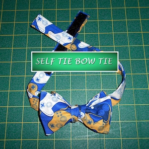 May include: A self-tie bow tie with a blue, white, and gold controller pattern. The bow tie has a hook and loop closure. The text "SELF TIE BOW TIE" is displayed on the image.