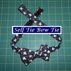 May include: A self-tie bow tie with a Halloween theme. The black fabric is patterned with white ghosts and orange jack-o'-lanterns. The bow tie has a hook and loop closure. The text "Self Tie Bow Tie" is in a blue box.