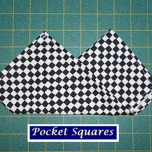 May include: A black and white checkered pocket square folded into a triangle shape. The fabric is a cotton blend and has a subtle sheen. The pocket square is on a green cutting mat with a grid pattern.
