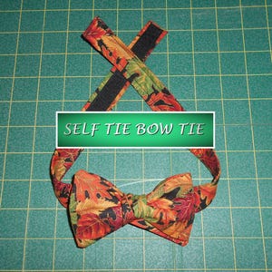 May include: A self-tie bow tie with a fall leaf pattern in shades of red, orange, green, and black. The bow tie is displayed on a green grid surface, with the text "SELF TIE BOW TIE" in a white font.