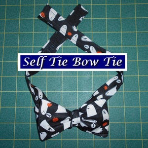 May include: A black self-tie bow tie with a Halloween-themed pattern. The fabric features white ghosts and orange pumpkins on a black background. The text "Self Tie Bow Tie" is displayed on a blue and white banner.