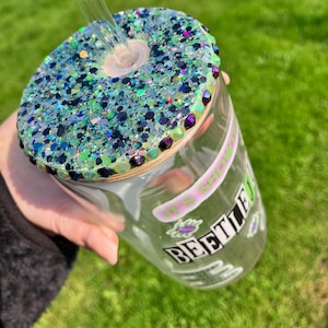 以下が含まれることがあります： キラキラ光る、ティールと紫色の蓋が付いた透明なガラスタンブラー。蓋の縁には小さな紫色の宝石が飾られています。「BEETLE」と「IT'S SHOWTIME」の文字がプリントされています。