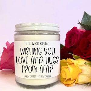 May include: White soy candle with a white lid and a label that reads "The Wick Club Wishing You Love and Hugs From Afar Handcrafted 8oz. Soy Candle".