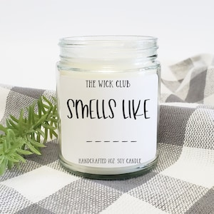 May include: A white soy candle in a clear glass jar with a white label that reads "The Wick Club" and "Smells Like" with a blank line for the scent name. The candle is on a white and gray checkered surface.