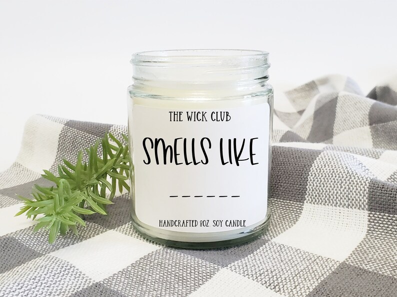May include: A white soy candle in a clear glass jar with a white label that reads "The Wick Club" and "Smells Like" with a blank line for the scent name. The candle is on a white and gray checkered surface.
