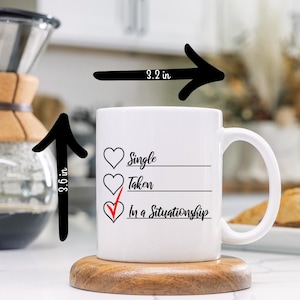 Puede incluir: Taza de cerámica blanca con un diseño en negro y rojo. La taza dice "Soltero, Casado, En una relación" con una marca de verificación junto a la última opción. La taza mide 9,1 cm de alto y 8,1 cm de ancho.