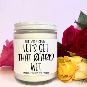 May include: A white soy candle with a black label that reads "The Wick Club Let's Get That Beard Wet Handcrafted 8oz Soy Candle". The candle is sitting on a white surface with a bouquet of red and yellow roses.