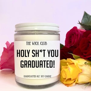 May include: A white soy candle with a black label that reads "THE WICK CLUB HOLY SH*T YOU GRADUATED! HANDCRAFTED 8OZ. SOY CANDLE". The candle is sitting on a white surface with a bouquet of red and yellow roses.