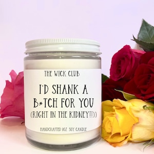 May include: A white candle jar with a black label that reads "THE WICK CLUB I'D SHANK A B*TCH FOR YOU (RIGHT IN THE KIDNEY!!)" HANDCRAFTED 8OZ. SOY CANDLE". The candle jar is sitting on a table with a bouquet of red and yellow roses.