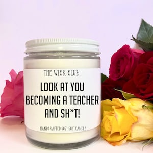 May include: A white soy candle with a black label that reads "THE WICK CLUB LOOK AT YOU BECOMING A TEACHER AND SH*T! HANDCRAFTED 8OZ. SOY CANDLE". The candle is surrounded by red and yellow roses.