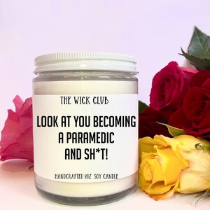 Peut inclure: Une bougie en soja blanche avec une étiquette noire qui indique "THE WICK CLUB LOOK AT YOU BECOMING A PARAMEDIC AND SH*T! HANDCRAFTED 8OZ. SOY CANDLE". La bougie est posée sur une surface blanche avec un bouquet de roses rouges et jaunes.