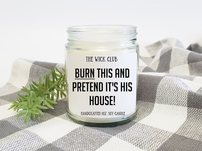 May include: A white soy candle in a clear glass jar with a black label that reads "THE WICK CLUB BURN THIS AND PRETEND IT'S HIS HOUSE! HANDCRAFTED 8OZ SOY CANDLE".