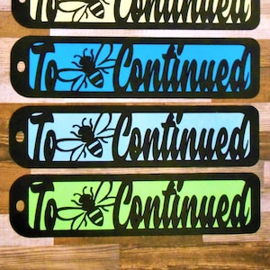 以下が含まれることがあります： 黒い文字で「To Be Continued」と書かれたブックマークが5つ。各ブックマークには、黒いシルエットの蜂と、ピンク、黄色、青、水色、緑の異なる色の背景があります。