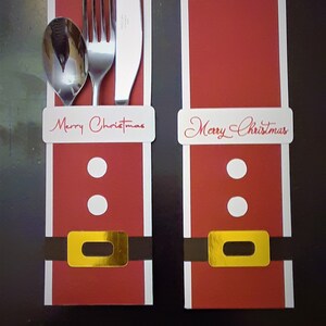 Puede incluir: Dos porta tarjetas de lugar de Navidad rojos y blancos con el texto "Feliz Navidad". Cada porta tarjetas tiene una hebilla dorada y dos círculos blancos. Un tenedor, un cuchillo y una cuchara de plata se colocan en el porta tarjetas de la izquierda.