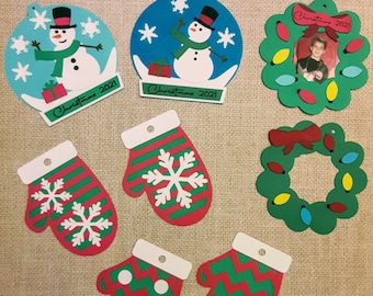 May include: Set of Christmas ornaments featuring a snowman in a snow globe, a wreath with lights, and mittens with snowflake designs. The ornaments are made of paper and have a red and green color scheme. The ornaments are labeled "Christmas 2021".