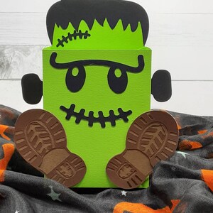 May include: Green Frankenstein monster-shaped box with black eyes, a stitched scar, and brown boot-shaped feet. The box is sitting on a black and orange fabric.