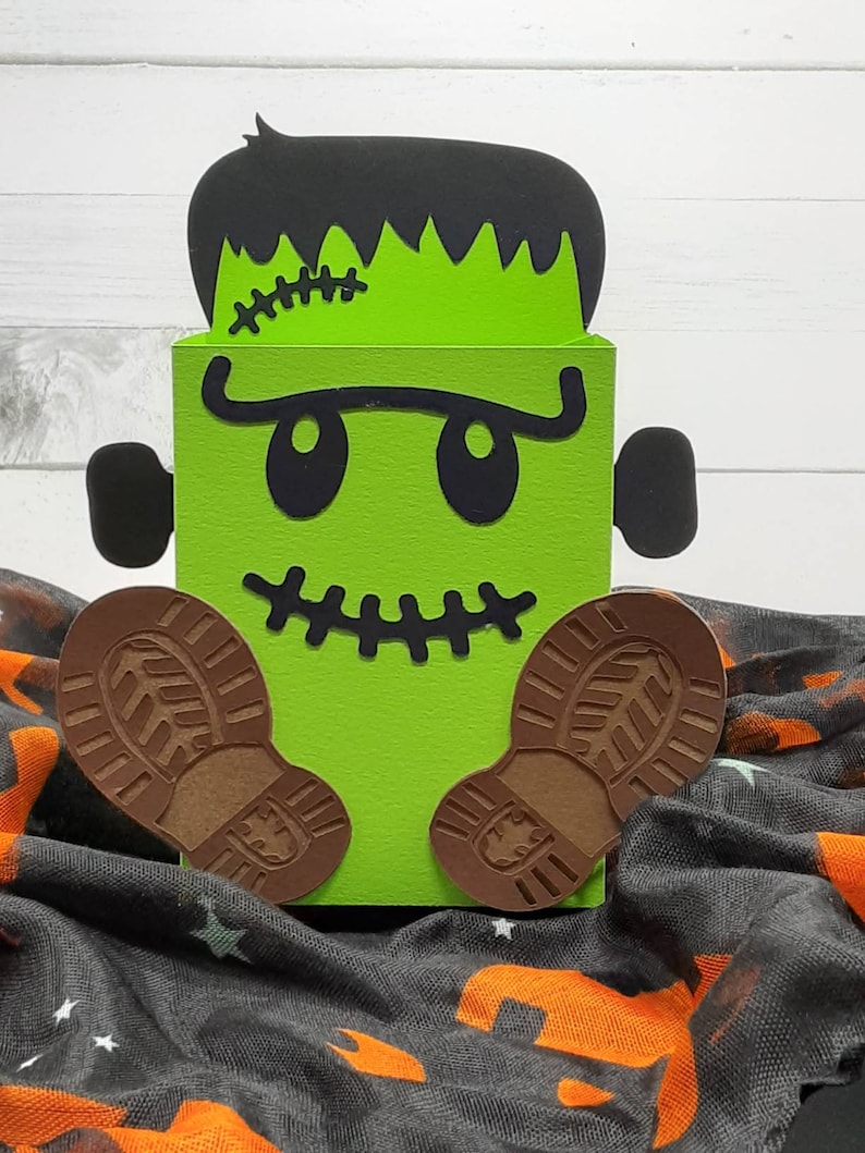 May include: Green Frankenstein monster-shaped box with black eyes, a stitched scar, and brown boot-shaped feet. The box is sitting on a black and orange fabric.