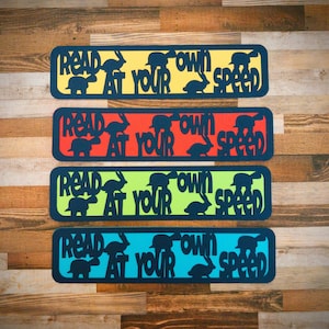 Può includere: Quattro cartelli rettangolari colorati con il testo "Leggi al tuo ritmo" in lettere nere. Ogni cartello ha un colore di sfondo diverso: giallo, rosso, verde e blu. I cartelli sono decorati con silhouette nere di animali, tra cui un coniglio, una tartaruga e un uccello.