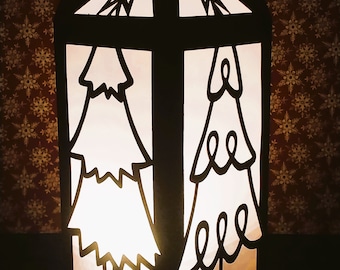 May include: A black paper lantern with a white silhouette of two Christmas trees and stars on the top. The lantern is lit from inside, casting a warm glow.