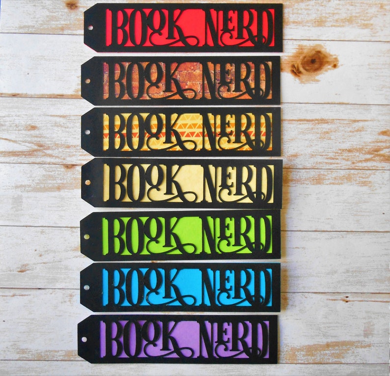 K&ouml;nnte beinhalten: Sieben schwarz-wei&szlig;e Lesezeichen mit dem Text "Book Nerd" in einer dekorativen Schriftart. Jedes Lesezeichen hat einen andersfarbigen Hintergrund: rot, braun, orange, gelb, gr&uuml;n, blau und lila.