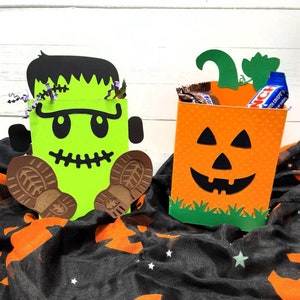May include: Two Halloween treat bags, one shaped like a green Frankenstein monster with brown boots and the other shaped like an orange pumpkin with a jack-o-lantern face. Both bags have green grass at the bottom.
