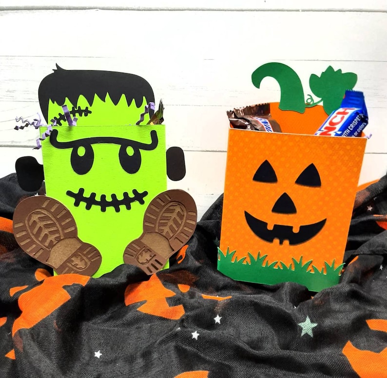 May include: Two Halloween treat bags, one shaped like a green Frankenstein monster with brown boots and the other shaped like an orange pumpkin with a jack-o-lantern face. Both bags have green grass at the bottom.