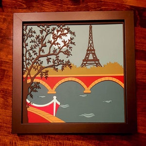 May include: A framed paper art piece depicting a stylized scene of the Eiffel Tower in Paris, France. The artwork features a red bridge with arches over a blue river, a tree with brown branches, and a full moon in the sky.