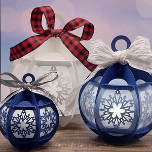 以下が含まれることがあります： 赤いチェックのリボンが付いた白い紙のクリスマスオーナメント、白いリボンが付いた青い紙のクリスマスオーナメント、銀色のリボンが付いた小さな青い紙のクリスマスオーナメントの3つ。それぞれのオーナメントには雪の結晶のデザインが施されています。