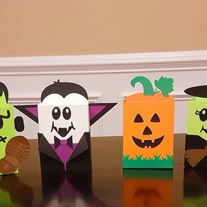 May include: Four Halloween-themed treat boxes, each featuring a different character: Frankenstein, Dracula, a pumpkin, and a witch. The boxes are made of cardboard and decorated with colourful paper and cutouts.