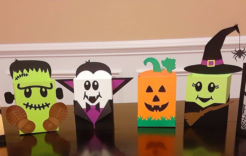 May include: Four Halloween-themed treat boxes, each featuring a different character: Frankenstein, Dracula, a pumpkin, and a witch. The boxes are made of cardboard and decorated with colourful paper and cutouts.