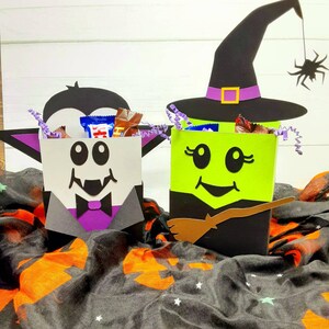 May include: Two Halloween treat boxes, one shaped like a vampire and the other shaped like a witch. The vampire box is black and white with a purple bow tie and fangs. The witch box is green with a black hat and a brown broom. Both boxes are filled with sweets.