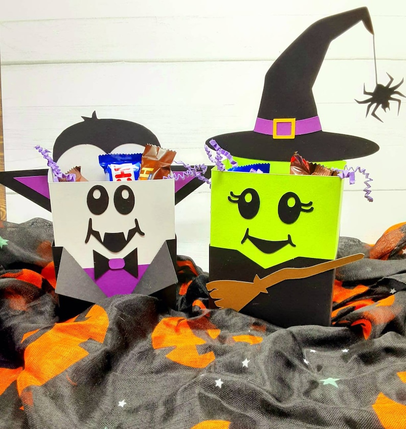 May include: Two Halloween treat boxes, one shaped like a vampire and the other shaped like a witch. The vampire box is black and white with a purple bow tie and fangs. The witch box is green with a black hat and a brown broom. Both boxes are filled with sweets.